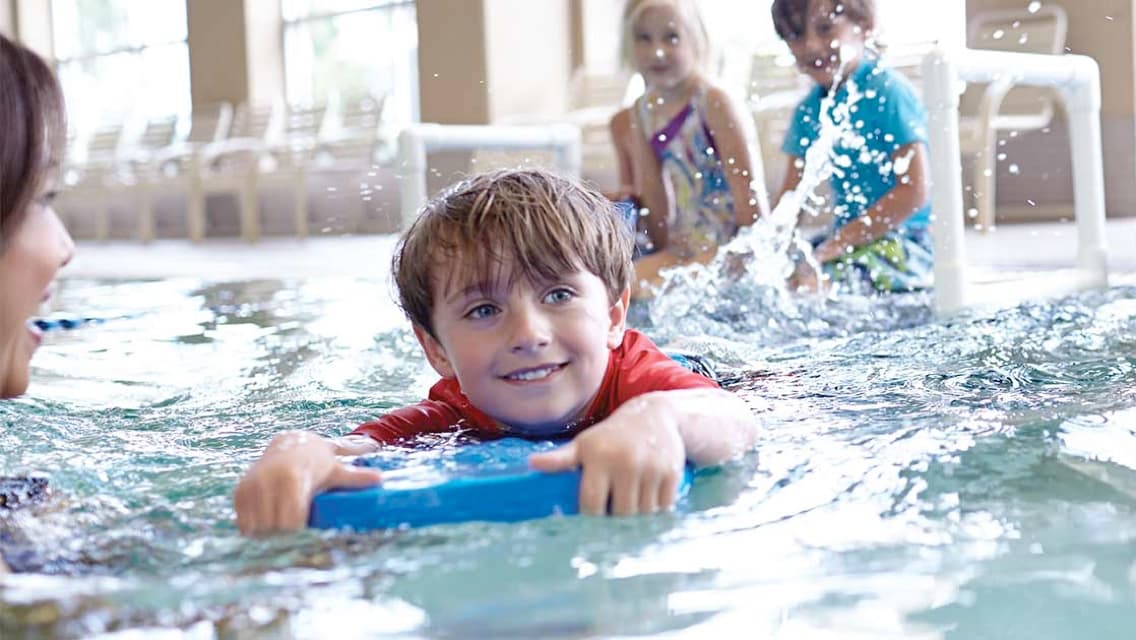 Young boy swimming in pool with kickboard alongside instructor with friends smiling while watching