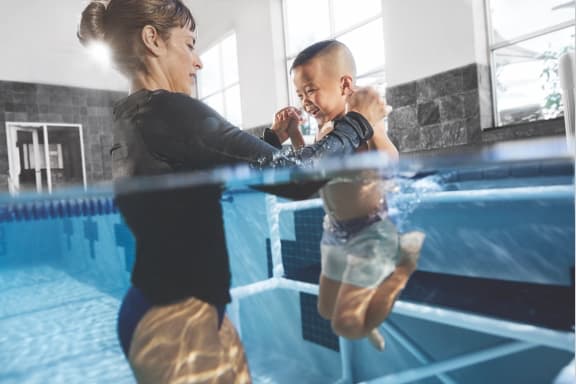 Swim instructor grabbing the hands of a smiling young boy learning how to swim